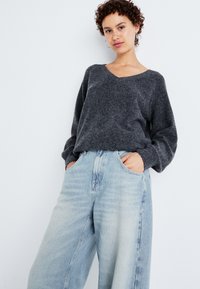 Maglione grigio a collo a V con maniche lunghe abbinato a jeans a gamba larga di lavaggio chiaro, dotati di tasche laterali e un fit rilassato.