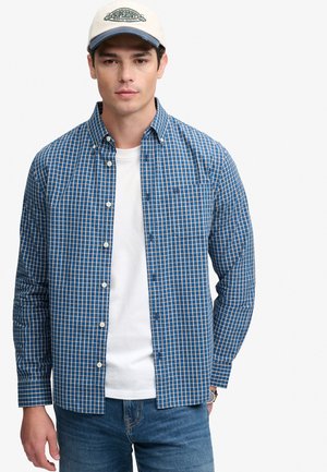 Mand iført en blå ternet skjorte over en hvid t-shirt, blå jeans, en beige kasket og et armbåndsur på venstre håndled.