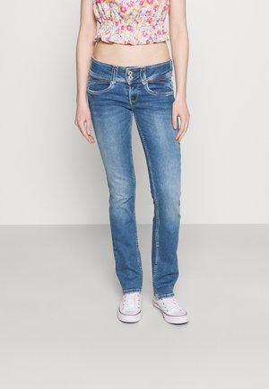 Kvinde iført falmede blå slimfit-jeans, blomsterprintet crop top og hvide sneakers, stående mod en ensfarvet lys baggrund.