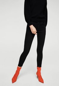 Jeans ajustados de color negro y una blusa negra de manga larga combinados con botas de tacón alto de tobillo de un brillante color naranja sobre un fondo neutro.