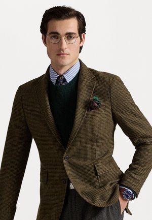 Braun karierter Blazer, über einem dunkelgrünen Pullover und einem hellblauen gestreiften Hemd getragen. Verfügt über runde Brillen und ein gemustertes Einstecktuch.