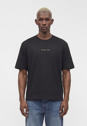 Uomo con capelli biondi corti che indossa una T-shirt nera Michael Kors e jeans blu, in piedi davanti a uno sfondo chiaro uniforme.