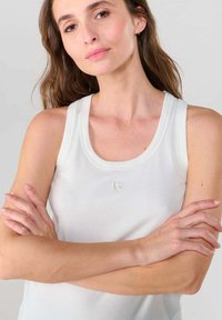 Witte geribbelde tanktop met een ronde hals en een klein logo op de borst. Model poseert met de armen gekruist voor een neutrale achtergrond.