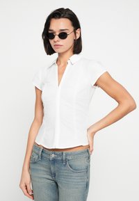 Άσπρο στενό κοντομάνικο πουκάμισο με γιακά και κουμπιά μπροστά. Το μοντέλο φοράει μαύρα στρογγυλά γυαλιά ηλίου και ανοιχτόχρωμα μπλε τζιν. Απλός σχεδιασμός.