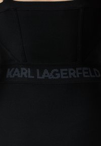 Črnen tekstilni kos z reberci, ki ima izrazito horizontalno logotipno pás z napisom "KARL LAGERFELD" v kontrastni barvi.