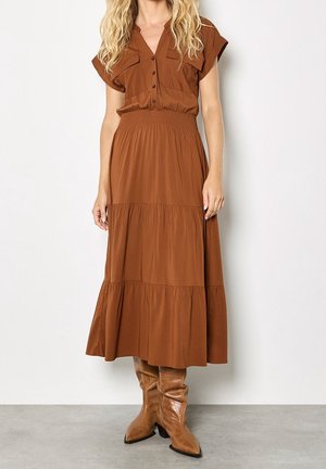 Femme portant une robe marron à boutons avec col en V, jupe à volants et bottes en cuir marron mi-mollet assorties, debout sur un sol gris.