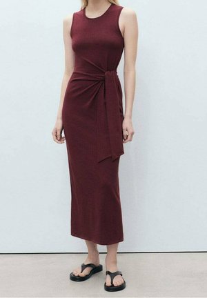 Woman wearing a sleeveless, ankle-length, burgundy dress with a side knot detail, paired with black platform sandals.
