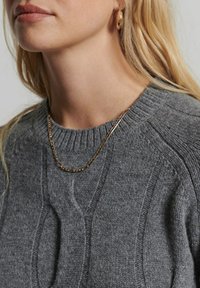 Gros plan d'une personne portant un pull gris en tricot, un collier chaîne en or, et des boucles d'oreilles anneaux en or assorties, avec des cheveux blonds.