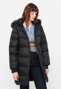 Zwarte pufferjas met een zachte bontafwerking aan de capuchon, ritssluiting aan de voorkant en een doorgestikt patroon. Draagt over een grijze top en een blauwe cardigan.