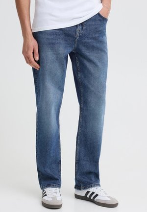 Homme portant un jean bleu, une chemise blanche et des baskets blanches à rayures noires, debout avec une main dans la poche sur un fond uni.