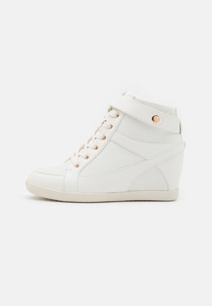 Hvid højt skiltesneaker med snørelukning foran og ankelrem med gylden knap på ensfarvet baggrund.
