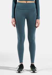 Rippelige, türkisfarbene Leggings aus dehnbarem Stoff; hoher Bund; dezente Markenkennzeichnung an der Seite; kombiniert mit schwarzen Sportschuhen.