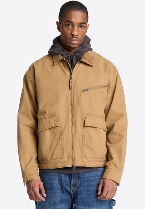 Mand iført en beige lynlåstrøje over en mørkegrå hættetrøje og blå jeans, stående med et neutralt ansigtsudtryk.