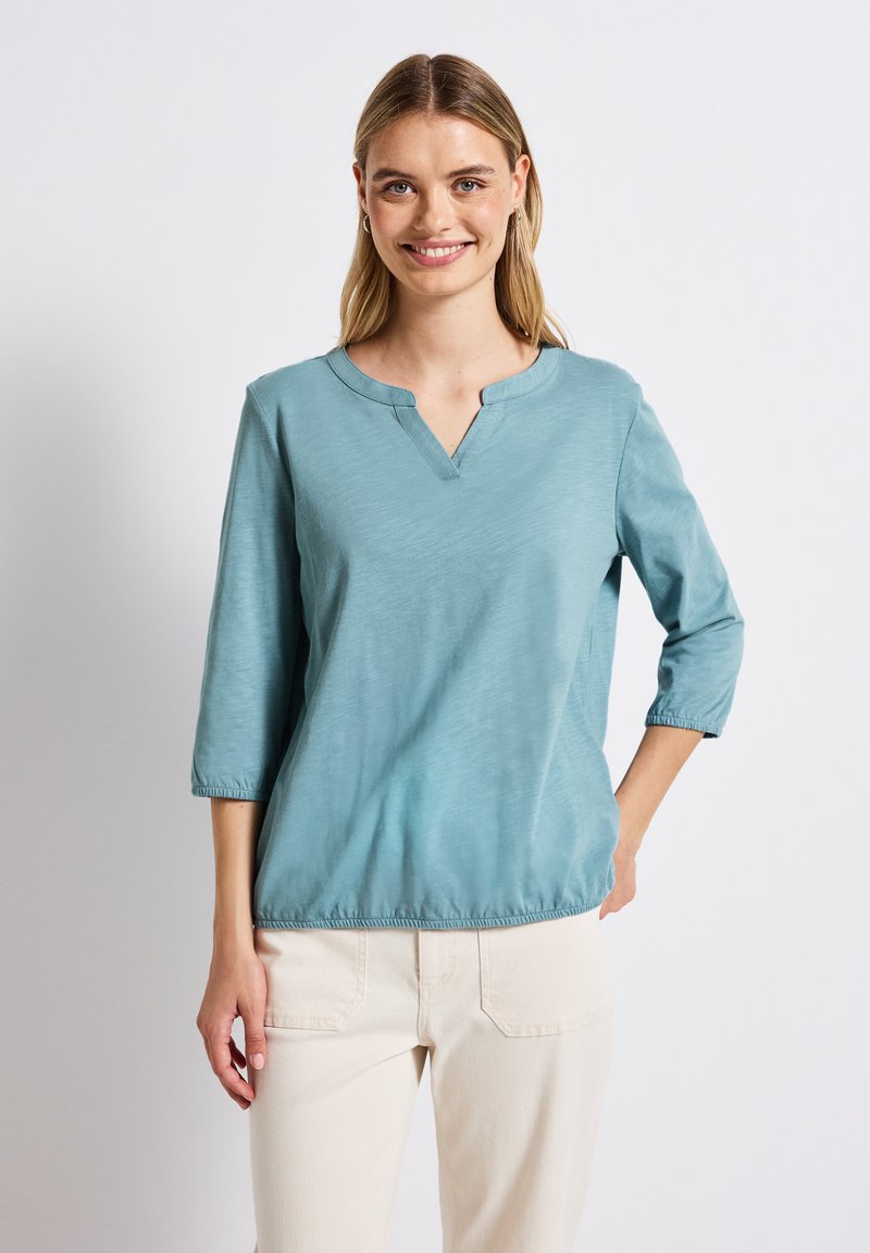 Teal toppi, jossa on kolme neljännestä hihaa, V-pääntie ja joustava helma, valmistettu pehmeästä kankaasta, jossa on hienovarainen tekstuuri ja rento istuvuus.