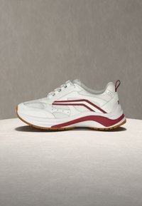 Λευκό αθλητικό παπούτσι με κόκκινες λεπτομέρειες, πλέγμα στα πλαϊνά μέρη και σόλα από καουτσούκ καφέ, τοποθετημένο σε ουδέτερη επιφάνεια πάνω σε απλό φόντο.