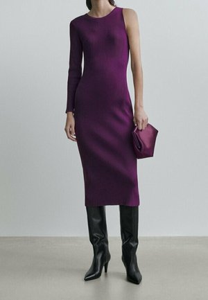 Femme portant une robe midi violette sans manches avec une manche longue, tenant une pochette violette assortie, accompagnée de bottes noires montantes jusqu'aux genoux.