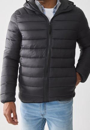 Homme portant une veste matelassée noire zippée avec une capuche par-dessus une chemise blanche et un jean bleu, une main dans la poche.