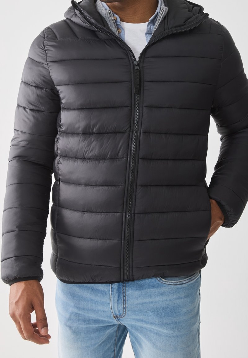Homme portant une veste matelassée noire zippée avec une capuche par-dessus une chemise blanche et un jean bleu, une main dans la poche.
