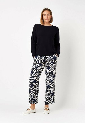 Donna in piedi con le mani in tasca che indossa una maglia nera a manica lunga, pantaloni blu navy e bianchi con motivo geometrico e scarpe bianche basse con fibbie.