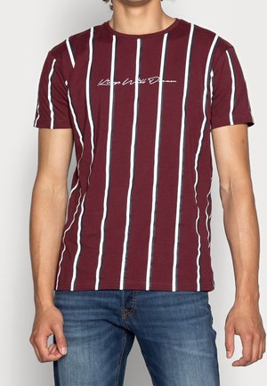 Mann trägt burgunderfarbenes T-Shirt mit kurzen Ärmeln, vertikalen weißen und schwarzen Streifen und kursivem Text, kombiniert mit blauen Denim-Jeans.