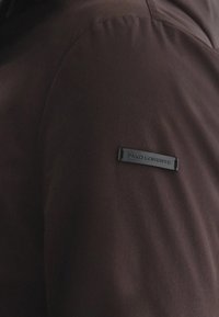 Brązowa kurtka o gładkiej fakturze z czarną metką z napisem "PAKO LORENTE" na ramieniu, prezentująca minimalistyczny design.