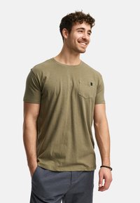 Camiseta de manga corta en verde oliva, hecha de algodón suave, con un bolsillo en el lado izquierdo del pecho que lleva una pequeña etiqueta negra.
