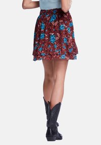 Jupe florale rouge avec des accents bleus, dotée d'un design à volants superposés. Le matériau semble léger. Associée à des bottes de cowboy noires.