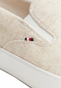 Scarpa slip-on beige con texture, pannello laterale elastico bianco e piccolo logo ricamato nero, bianco e rosso vicino alla suola.