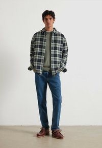 Camisa de cuadros azul marino y verde claro, superpuesta a un suéter verde texturizado. Combinada con jeans de mezclilla azul y zapatos marrones con cordones.