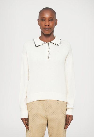 Maglione crema con colletto a righe nere, vestibilità rilassata, polsini e orlo a costine. Abbinato a pantaloni leggeri beige con un motivo a piccoli punti.