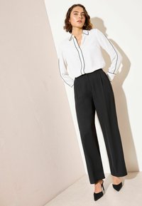 Biela košeľa na gombíky s čiernym lemovaním, v kombinácii s čiernymi, vysokým pásom a skladanými nohavicami. Modelka má na sebe čierne špicaté podpätky.