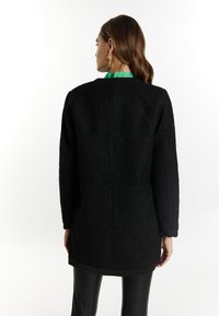 Cappotto nero a trama con taglio dritto, maniche lunghe e orlo arrotondato, indossato sopra pantaloni neri aderenti. Dettaglio verde sul colletto interno visibile.