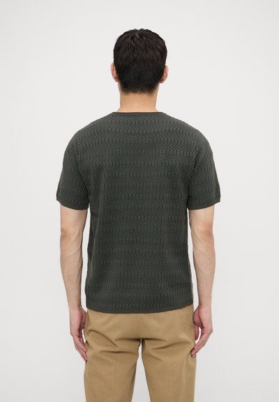 Kortærmet, mørkegrøn strikket t-shirt med et tekstureret zigzag-mønster. Rund hals og en afslappet pasform, parret med tanfarvede bukser.