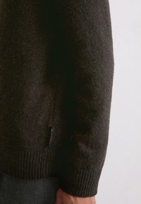 Dunkelbrauner Strickpullover mit strukturiertem Finish, ausgestattet mit gerippten Bündchen und Saum sowie einem kleinen Markenetikett an der unteren Seite.