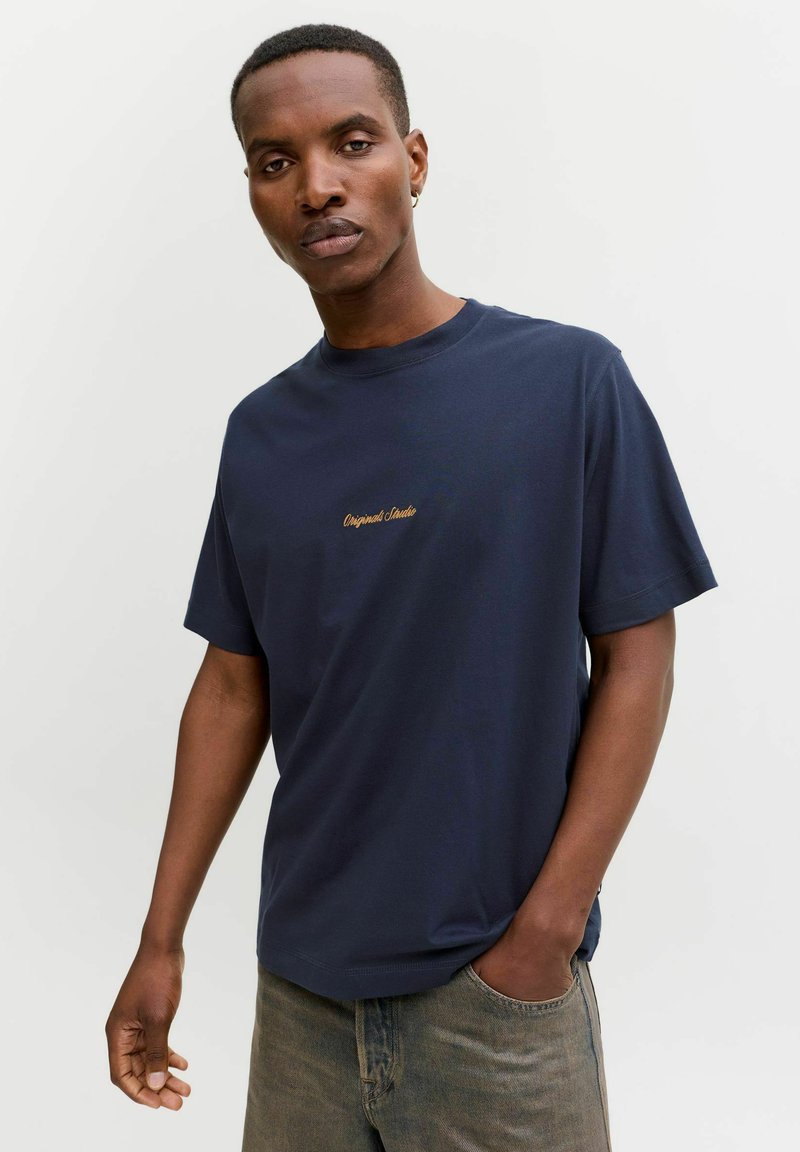 Tumesinine puuvillane T-särk lühikeste varrukatega, ümmargune kaelus ja diskreetne kuldne broderiga tekst rinnal. Seljas hallid teksapüksid.