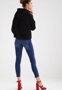 Sudadera negra con un corte holgado y capucha, combinada con jeans skinny azules y sandalias de tacón rojas. Diseño simple y moderno.