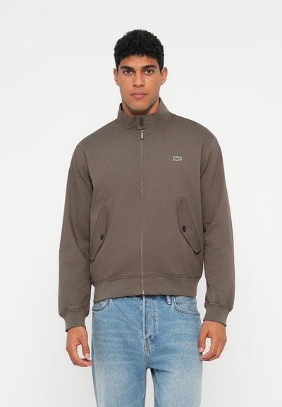 Homme debout portant une veste brune zippée avec des poches boutonnées et un jean bleu clair, devant un fond blanc uni.
