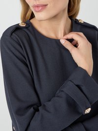 Μπλε ναυτικού τοπ με ριγέ υφή, στρογγυλή λαιμόκοψη και χρυσά κουμπιά στα ώ shoulder ασ; το μοντέλο κρατά απαλά το ύφασμα στη λαιμόκοψη.