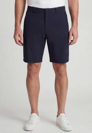 Homme portant un short cargo bleu marine avec des poches latérales et des baskets blanches, debout devant un fond clair uni.