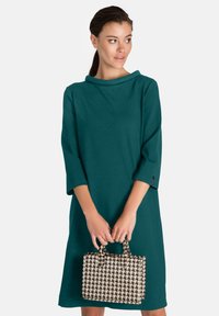 Rochie teal în formă de A, cu mâneci trei sferturi, decolteu larg și un material ușor. Ținând o mică geantă de mână cu model houndstooth.