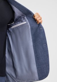 Blazer blu navy con un esterno testurizzato e una fodera grigia liscia, caratterizzato da una tasca interna e dettagli di cucitura. Una mano tiene il flap aperto.