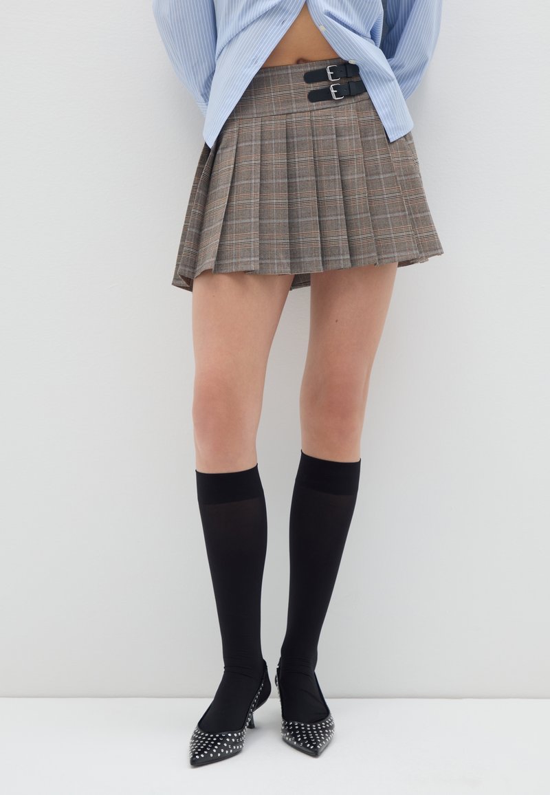 Braun kariierter, gefälteter Rock mit doppelten schwarzen Schnallenakzenten, kombiniert mit knielangen schwarzen Socken und spitz zulaufenden Schuhen, die mit Nieten verziert sind.