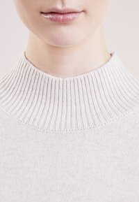 Pull en maille gris clair avec un col large en côtes, texture douce et design minimaliste. Motif délicat sur le tissu.