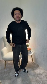 Homme aux cheveux bouclés portant un sweat-shirt noir, un jean foncé et des baskets, debout dans une pièce avec un fauteuil crème et une table d'appoint avec des livres.