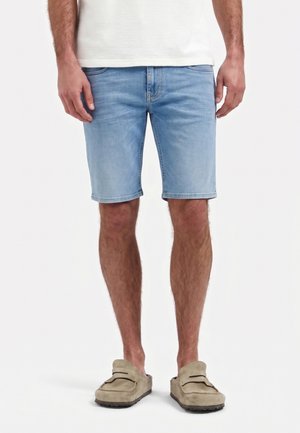 Mann trägt hellblaue Jeansshorts, beige Slipper und ein weißes Hemd, steht vor einem schlichten weißen Hintergrund.