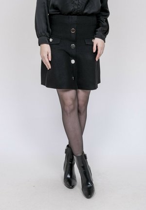 Femme portant une jupe noire boutonnée sur le devant, des collants noirs transparents, des bottines noires et un chemisier noir à manches longues, debout, jambes croisées.