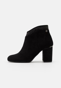 Botines de ante negros con punta redonda, tacón ancho tipo bloque y ribete con tachuelas alrededor del tobillo. Presentan un logotipo de acento plateado en el lateral.