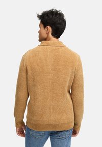 Beżowy sweter z prążkowanej tkaniny, z kołnierzykiem, dopasowany krój i teksturowany materiał, w połączeniu z niebieskimi dżinsami. Prosty design, bez widocznych wzorów czy akcentów.