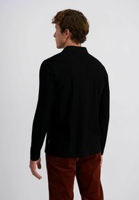 Man met kort bruin haar, gekleed in een zwart longsleeve shirt en roestkleurige corduroy broek, met zijn rug naar de camera gekeerd tegen een effen achtergrond.