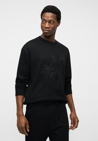 Νέος άνδρας που φοράει μαύρο φούτερ με ανάγλυφο σχέδιο φοίνικα, κοιτάζει προς τα δεξιά, στέκεται μπροστά σε απλό ανοιχτό φόντο.